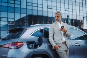 Geschäftsmann hält sein Smartphone in der Hand, während er sein Auto an einer Ladestation für Elektrofahrzeuge auflädt